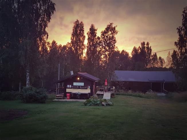 En stuga i en lantlig miljö omgiven av en grön gräsmatta och trädgårdsbyggnader, fotograferad vid solnedgång eller soluppgång.