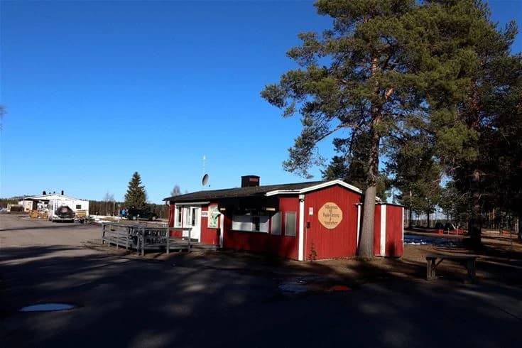 Bild av Pajala Camping Route 99 visar en asfalterad väg omgiven av grönska. En skuggad viloplats med en stolpe nära en rastplats och en byggnad som liknar ett värdshus syns i bakgrunden.