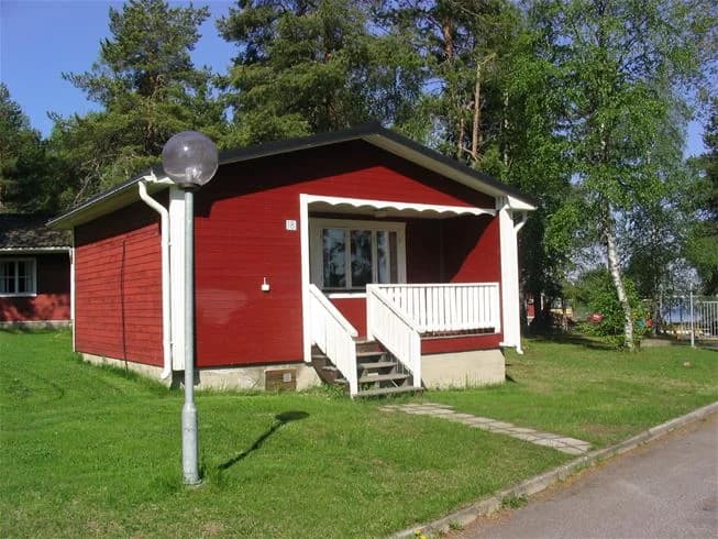 En röd stuga med vita knutar och tegeltak, omgiven av en välskött gräsmatta. Stugan har en liten veranda och en vit ytterdörr. Perfekt boende för besökare längs Pajala Camping Route 99.