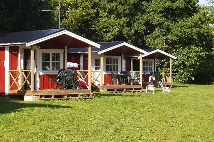 En campingstuga vid Sandbybadets camping omgiven av en grön gräsmatta. Framför stugan finns utemöbler i skuggan, perfekta för avkoppling.
