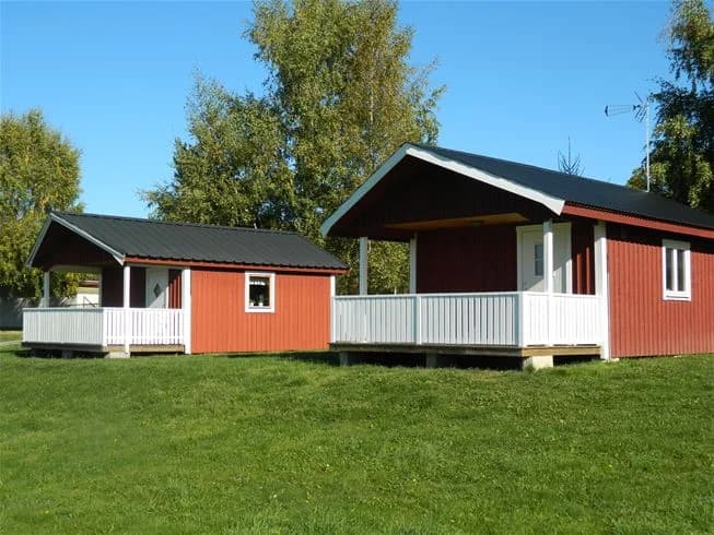 En lantlig stuga med tegeltak, omgiven av en välskött gräsmatta och trädgård med skuggiga ytor, typiska byggnader som en bod syns i bakgrunden.