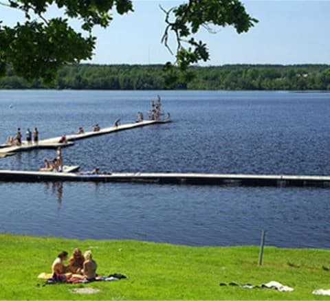 En somrig vy över Östanå camping med glittrande vatten och skogsklädda stränder. I förgrunden syns campingens strandnära brygga där människor kopplar av och njuter av sin semester. Perfekt för rekreation och friluftsliv.