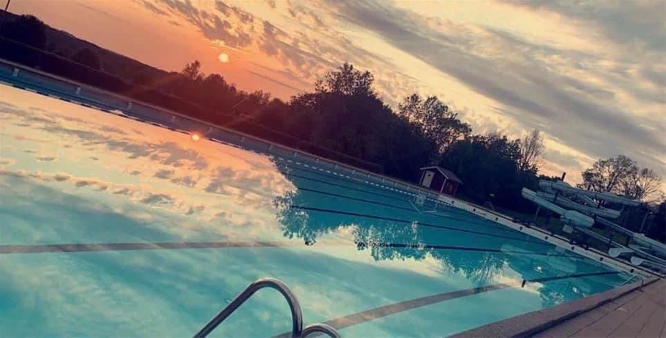 En upplyst swimmingpool i en resort vid Sannabadet under skymningen, omgiven av avslappnade besökare som njuter av en kväll vid poolen med en färgglad solnedgång i bakgrunden.