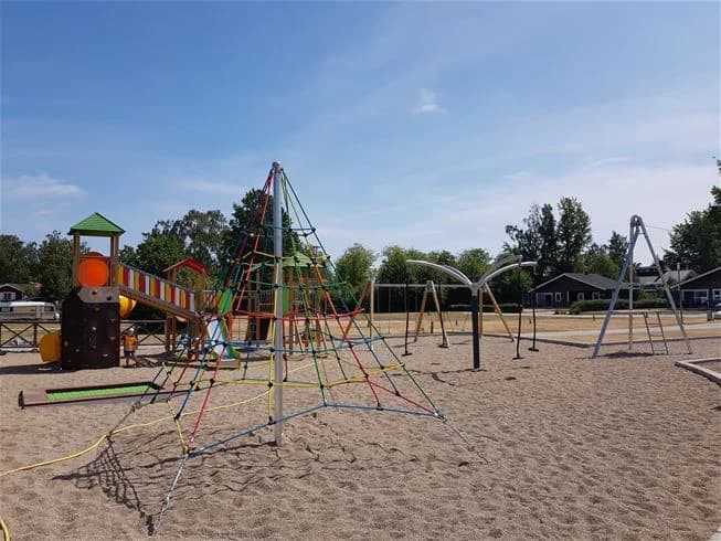 En campingplats med utsikt över en lekplats som har en rutschkana. Marken är omgiven av grönska och skugga, och i bakgrunden syns bebyggelse. Himlen är blå, vilket ger en avslappnad och inbjudande atmosfär.