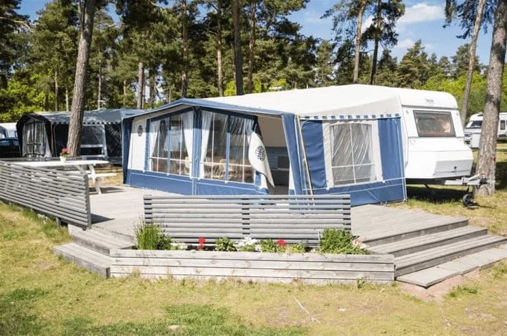 En skuggig campingplats på Kalmar Camping - Rafshagsudden med husvagn, markis och träaltan, omgiven av grönska.