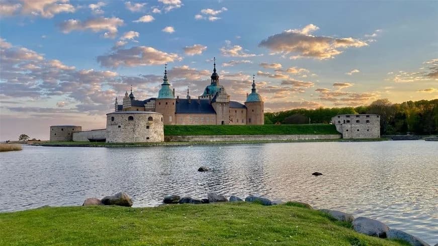 En kastelliknande byggnad belägen vid vatten med omgivande medeltida arkitektur och molntäckt himmel, en populär turistattraktion i Kalmar.