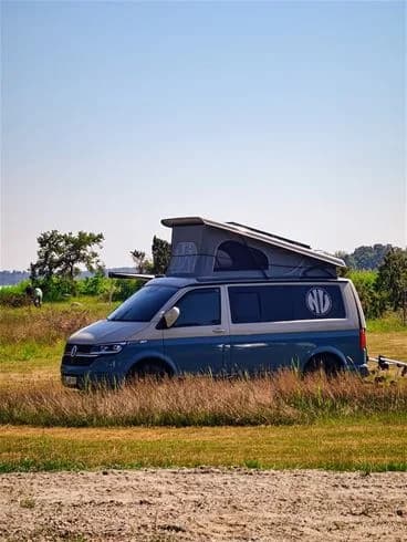 En vit Volkswagen husbil är parkerad vid Kalmar Camping - Rafshagsudden, omgiven av grönska.