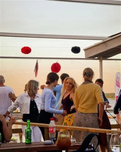 En grupp människor på Kalmar Camping - Rafshagsudden njuter av en solig dag vid vattnet. Flera av dem bär solglasögon och ser ut att ha roligt. Det är en livlig atmosfär med glada människor som deltar i olika aktiviteter.