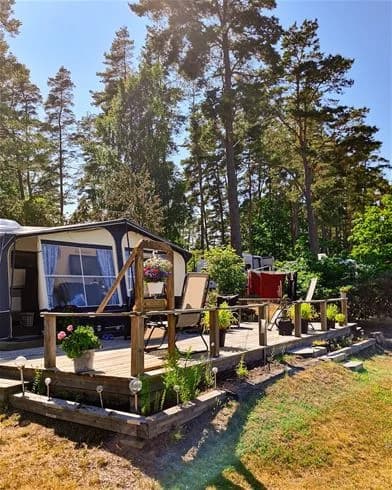 En lummig trädgård med stolar och bord i skuggan. En cykel står parkerad intill en husvagn i bakgrunden, omgiven av grönt gräs och träd, vilket ger en inbjudande känsla av friluftsliv och avkoppling.