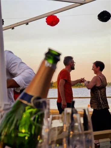 En grupp människor njuter av en ledig stund på Kalmar Camping - Rafshagsudden. På ett bord står flaskor och glas, och några personer bär solglasögon under en solig dag. Stämningen är avslappnad och festlig.