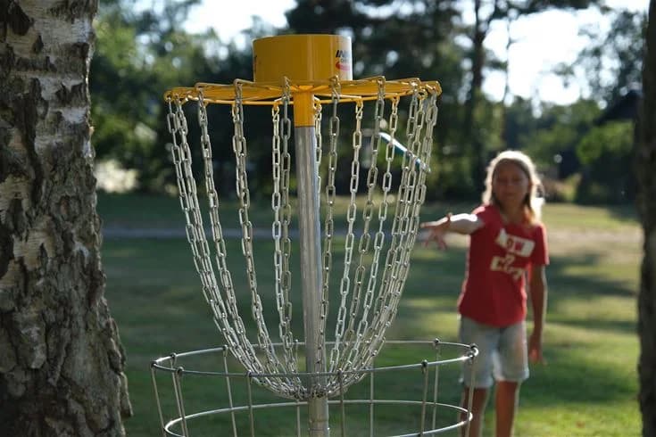 En grupp människor spelar discgolf i en skogsdunge på Gullbrannagården under en solig dag. Träden omringar området och skapar en naturskön plats för rekreation och lek.
