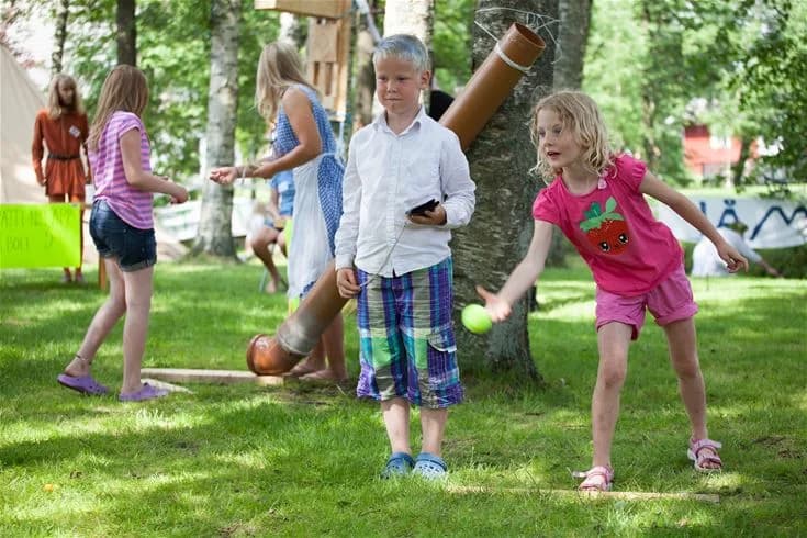 En grupp barn i shorts njuter av sommarlek på Gullbrannagården. De skrattar och har roligt i en grönskande utomhusmiljö fylld av glädje och gemenskap.