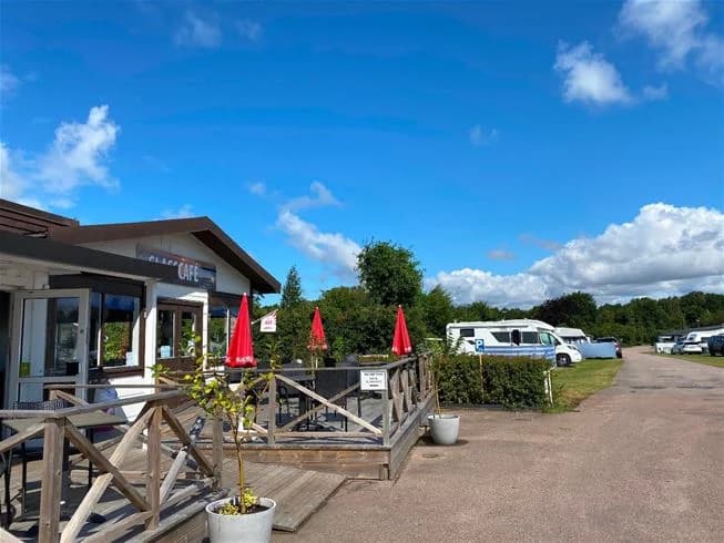 Bilden visar en parkeringsplats vid Vilshärads camping med flera bilar, inklusive familjebilar och SUV:ar. I bakgrunden syns en restaurang med uteservering och picknickbord, omgiven av gröna omgivningar.