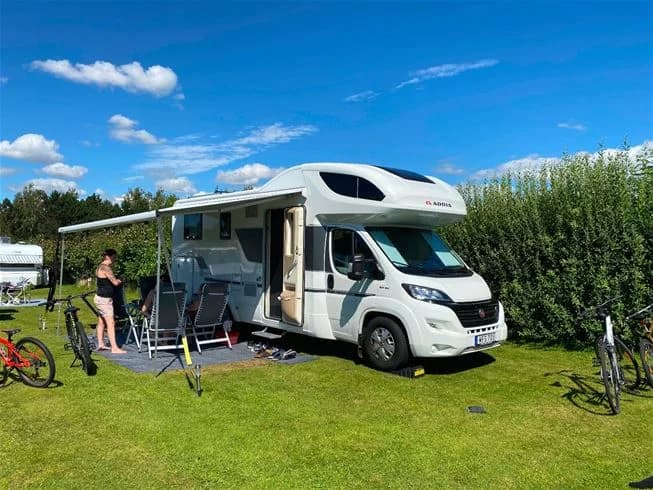 En husvagn står uppställd på Vilshärads camping omgiven av grönska. Framför husvagnen syns en person som kopplar av och några cyklar står parkerade, vilket ger en känsla av fritid och rekreation.