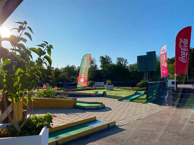 En översiktsbild över Vilshärads camping med fokus på en minigolfbana med konstgräs. I bakgrunden syns campingens logotyp och reklamskyltar. Området är omgivet av träd som ger skugga och det finns olika rekreationsytor för besökare.