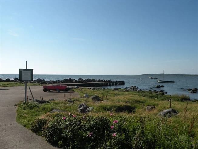 En pittoresk kustlinje vid Vilshärads camping med ett glittrande hav och en lugn strand. En båt syns i fjärran och en friluftsbänk står vid strandkanten, med en vik i bakgrunden.