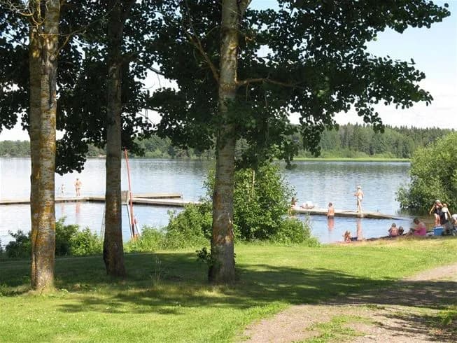 Bild av Falkudden Camping, Café och Stugby. I förgrunden syns ett rekreationsområde med gröna träd och växter runt en sjö eller damm. Området inbjuder till avkoppling och friluftsliv, med vatten och lekfull natur i bakgrunden.