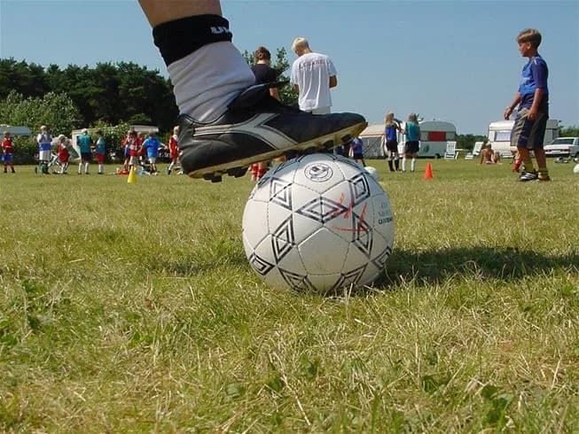 En grupp människor spelar fotboll på en grön gräsplan vid Gullbrannagården. I förgrunden syns en fotboll i rörelse och spelare i full rörelse med varierad sportutrustning._soligt väder och blå himmel ger en inbjudande atmosfär för utomhusaktiviteter.