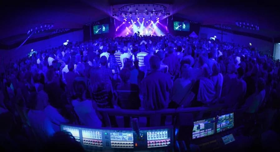 En folksamling njuter av en musikevent inomhus, med livfull belysning och skärmar som visar uppträdanden på Gullbrannagården. Publiken ser ut att ha roligt medan de deltar i evenemanget.