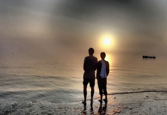 En grupp människor står vid strandkanten i skymningen vid Stenåsa stugor & camping. Solnedgången målar himlen i varma färger medan havet breder ut sig vid horisonten.