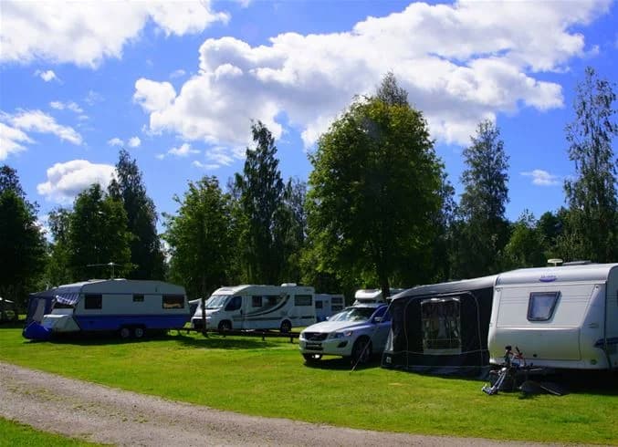 En grönskande campingplats med uppställda husbilar och husvagnar, omgivna av träd och öppna gräsytor, vilket skapar en lugn och naturnära atmosfär.