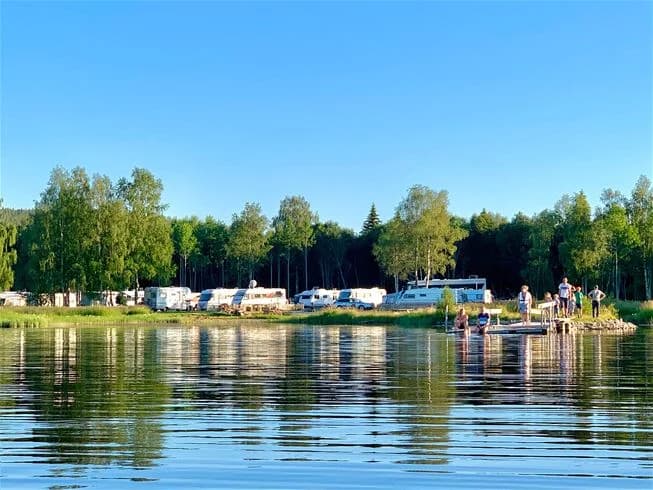 En somrig scen vid Överhörnäs camping, med en sjö eller vattendrag där en båt flyter. Grönskande stränder runt vattnet och en avslappnad atmosfär för rekreation.