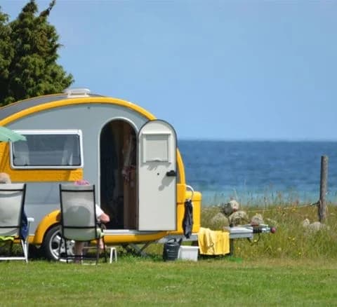 En campingplats med flera husbilar och husvagnar parkerade på gräs- och grusunderlag, omgiven av skog.