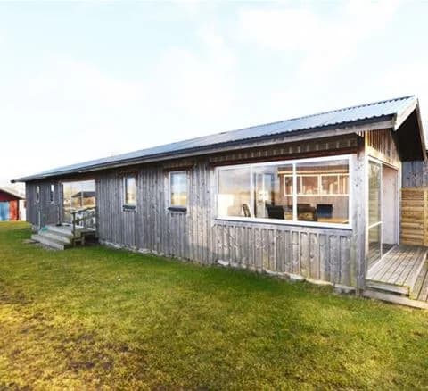 En trästuga med rött tak i en lantlig miljö med gräs och träd runtomkring. En dörr syns på stugans sida. Perfekt för besökare som söker avkoppling i Wikegårds semesterby & ställplatscamping.