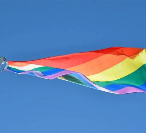 En campingplats med klart blå himmel och en färgglad regnbåge över en flagga som vajar i vinden. Solsken sprider ett gyllene sken över området.