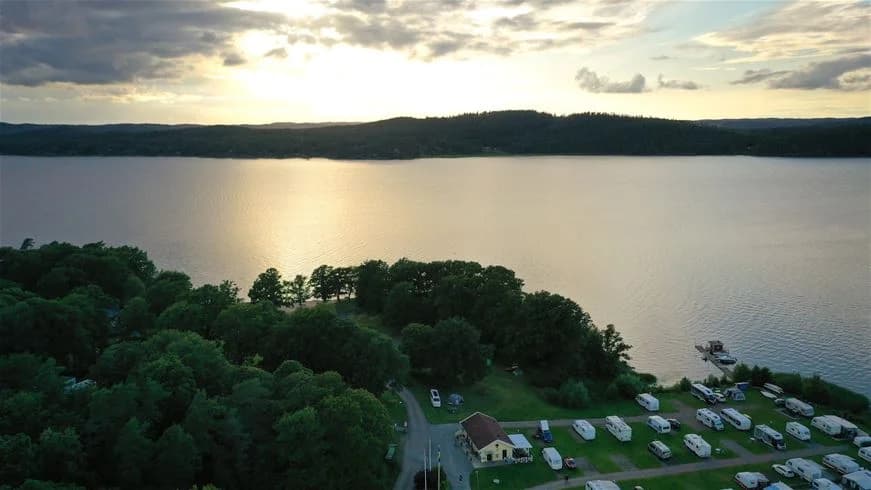 En naturskön vy över en lugn sjö med gröna träd längs strandkanten, omgiven av ett kuperat landskap vid Lövekulle camping.