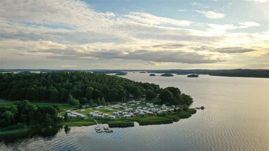 En naturskön campingplats vid en vattenkropp, omgiven av grönskande skogar och med en strandlinje som erbjuder utsikt över ett stilla vatten. Perfekt för friluftsliv och avkoppling i en idyllisk miljö.