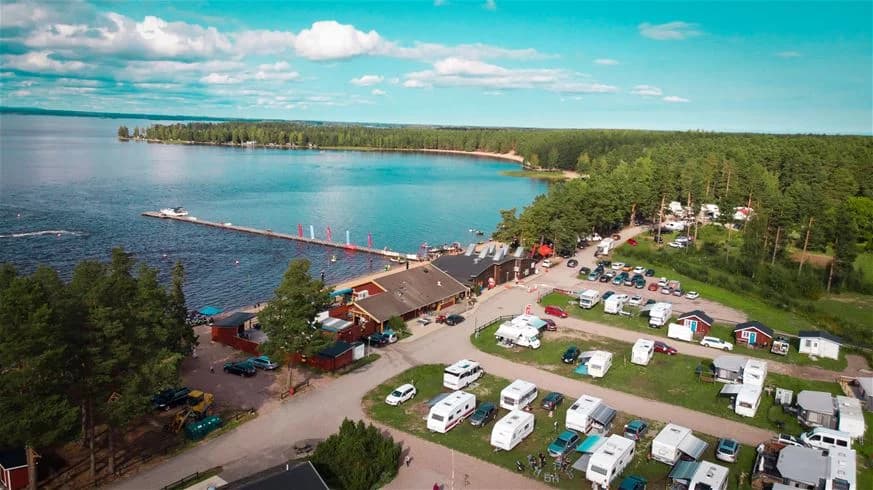 Luftvy över Årsunda Strandbad med sandstrand och brygga vid en sjö, omgiven av grönskande träd och semesteranläggningar.