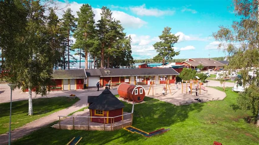 En solig dag vid Årsunda Strandbad, med gröna gräsmattor, lekplatser för barn och solskyddande träd i en avkopplande parkmiljö nära vattnet.