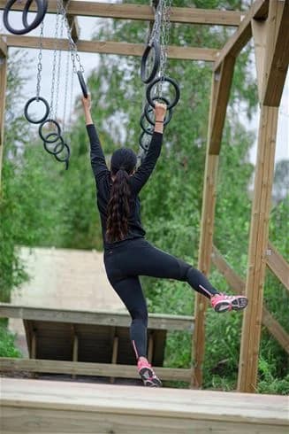 En person som njuter av utomhusaktiviteter vid Årsunda Strandbad. Balanserar på en repkonstruktion nära en lekplats, omgiven av grönska och soligt väder. Perfekt för rekreation och fysisk aktivitet.