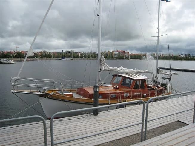 Vy över Västra Kajen Camping med flera båtar förtöjda i en småbåtshamn, omgivna av ett lugnt vatten och segelbåtsmaster som sträcker sig mot himlen.