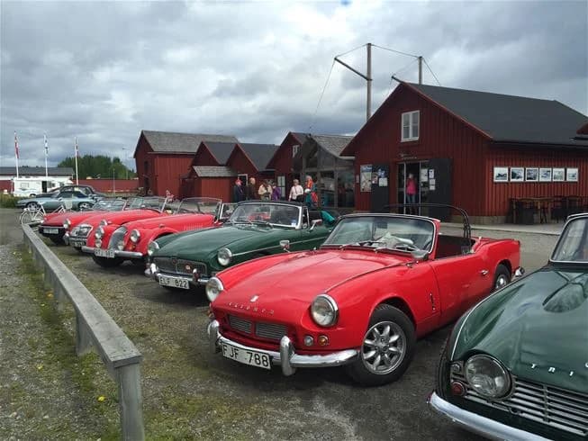 En veteranbil parkerad på Västra Kajen Camping, omgiven av grönska och andra bilar, med blå himmel i bakgrunden.