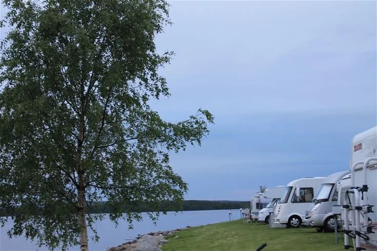 En campingplats med husbilar och husvagnar parkerade i rad vid västra kajen, omgivna av grönska och träd.