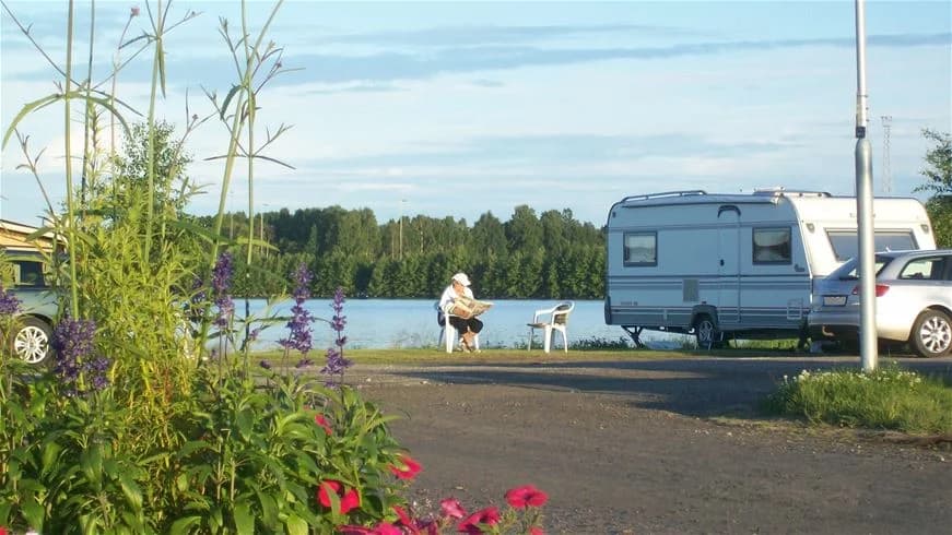 En husbil och en husvagn står parkerade på en grön och lummig campingplats vid Västra Kajen Camping. I bakgrunden syns fler träd och en klar himmel.