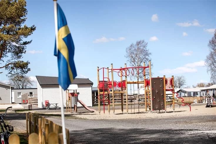 En lekplats med gungor och rutschkanor på Bingsmarkens camping. Solskydd över lekområdet, omgivet av barn som leker och träd som ger skugga.