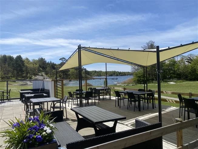 En bild av en campingplats vid Järnavik med utemöbler under ett solskydd, inklusive stolar och ett bord i förgrunden samt en baldakin och markis som ger skugga.