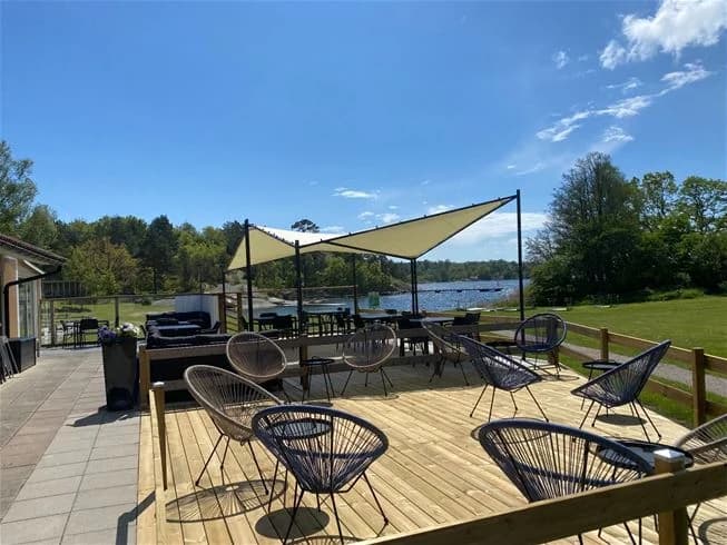 Bild av Järnaviks camping som visar en uteplats med trämöbler inklusive bord och stolar, beläget under skuggiga träd. I bakgrunden syns en grönskande parkmiljö som ger en inbjudande atmosfär för besökarna.
