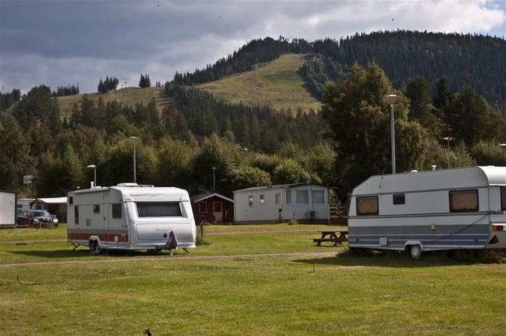 En naturskön campingplats med husvagn uppställd på grön gräsyta. Träd och öppna fält omger området, vilket ger en lantlig och avkopplande miljö.