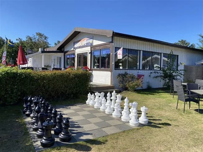 En grön gräsmatta med skuggade områden intill Vilshärads camping, omgiven av välskötta landskap och parkmiljö. I bakgrunden syns byggnader som kan vara ett motell eller restaurang.