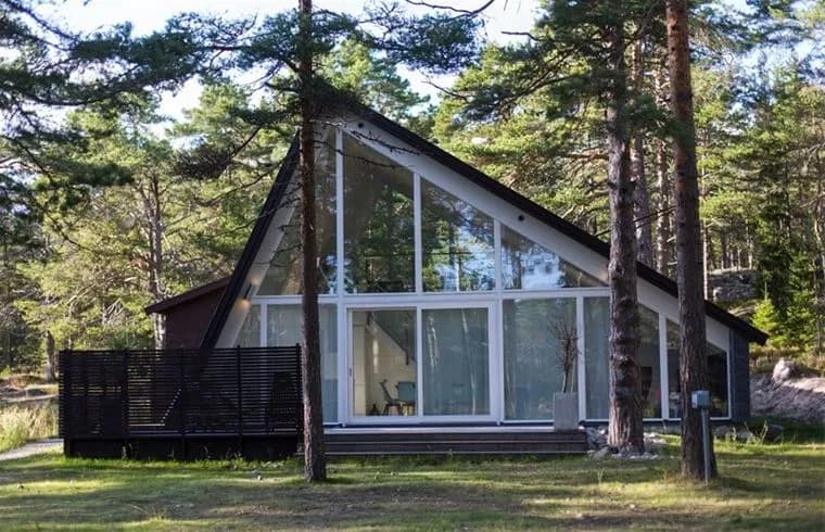 En vacker stuga med träpaneler omgiven av höga barrträd vid Hölick Havsresort. Stugan har ett rustikt utseende och står i skuggan av soliga träd, vilket skapar en harmonisk atmosfär.