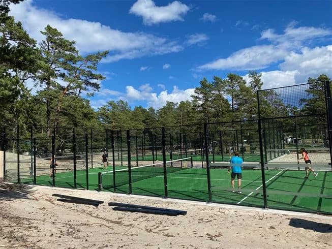 Alt-text: "En bild från Hölick Havsresort visar en fritidsanläggning med fokus på rekreation och sport. I förgrunden syns ett nät och staket, vilket antyder en sportaktivitet som racketspel. Området är omgivet av gröna träd och ger en känsla av avkoppling och nöje."