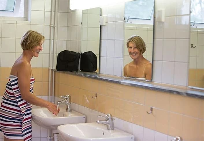 En blond person står vid en badrumsspegel på Borrbystrand Camping, med en handduk och ett handfat i förgrunden.