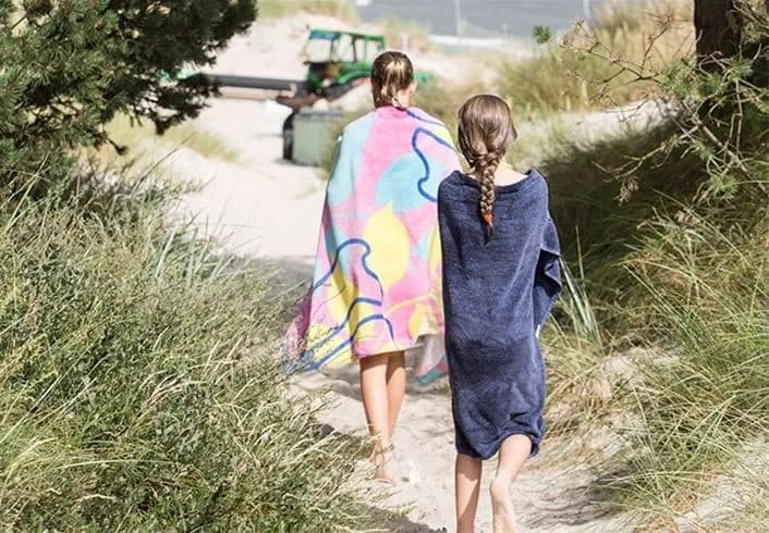 En grupp människor, barfota och iklädda badkläder och handdukar, njuter av en solig dag på stranden vid Borrbystrand camping. Sanden är gyllene och havet syns i bakgrunden, vilket skapar en avkopplande och naturnära atmosfär.