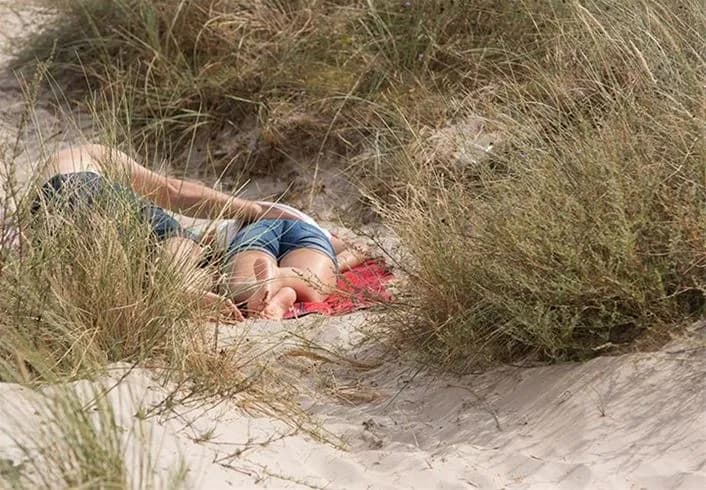 En solig sommardag på Borrbystrand camping med människor som solar och njuter av stranden. Sanddyner kantade av buskvegetation syns i bakgrunden.