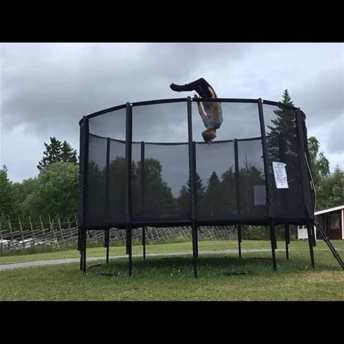 En bild av en campingplats på Sandvikens Camping och Stugby, med en stor trampolin omgett av grönska. Trampolinen är utrustad med ett skyddsnät och ligger under skuggiga träd, vilket skapar en avslappnad och trevlig miljö för rekreation.