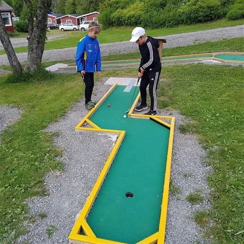 Bild av Sandvikens Camping och Stugby som visar en grönskande minigolfbana. Personer som spelar minigolf syns, omgivna av träd och gräs, vilket skapar en atmosfär av avkopplande fritidsaktivitet i naturen.